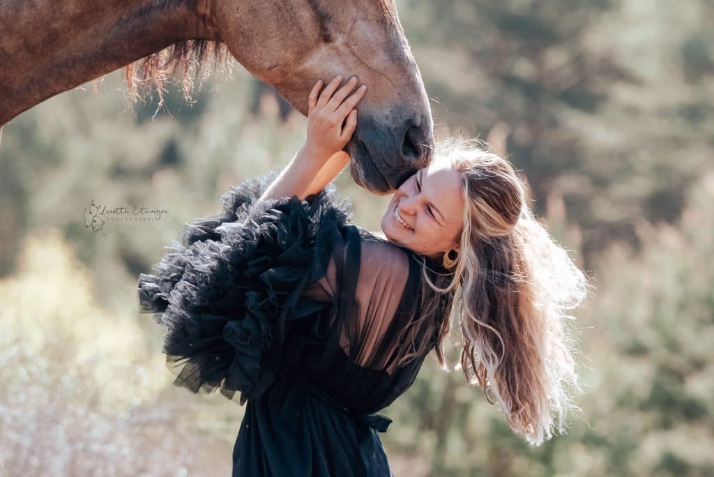 Eine Frau im schwarzen Kleid wird vom Pferd mit der Nase an die Wange gestupst