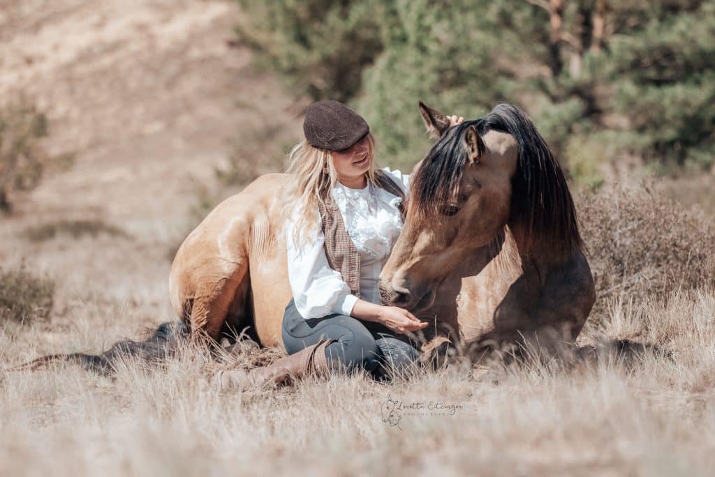 Eine Frau mit einem braunen Pferd wo das Pferd am boden liegt und die Frau daben sitzt.