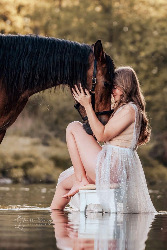 Eine Frau sitzt auf einem Stuhl im Wasser und ein braunes Pferd hält seinen Kopf an ihren.