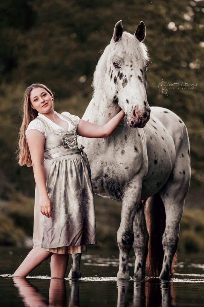Eine Frau steht im Wasser und ein Pferd (Schimmel) steht neben ihr.