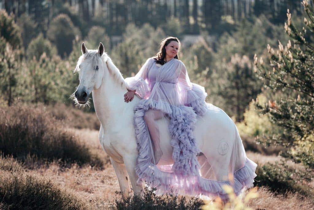 Eine Frau in Lila Kleid sitzt auf einem weißen Pferd