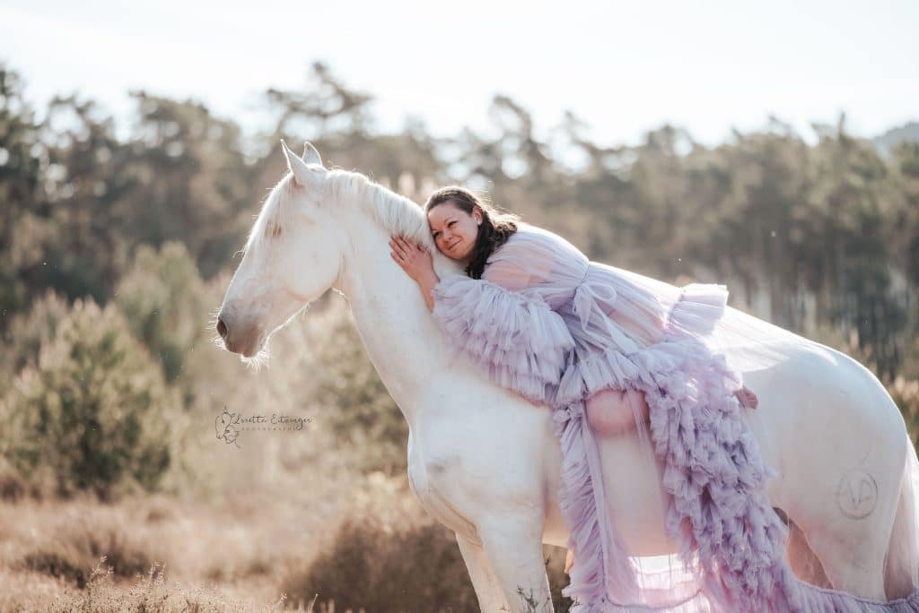 Eine Frau in einem Violetten langem Kleid sitzt auf einem weißen Pferd