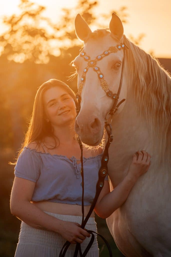 Eine Frau steht neben einem weißen Pferd welches einen Halfter mit Schmuck trägt.