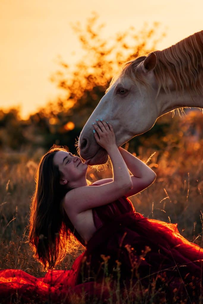 Eine Frau mit einem Pferd kniet am Boden und das Pferd leckt ihr ins Gesicht.